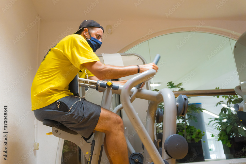 Caucasian young man with face mask on a gym machine performing exercises, gym with little vacuum, new normal, social distance, covi-19, coronavirus, pandemic