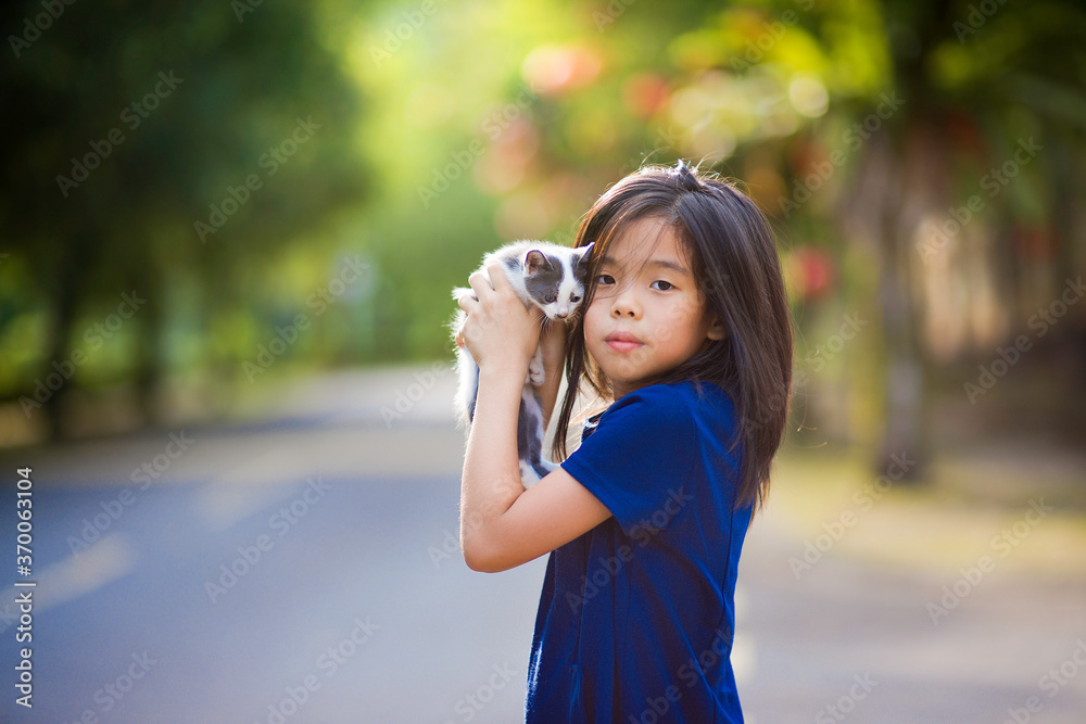 Asian young girl holding a cat kitten in the park under the sun. Pet and domestic animal lover. Happy child in nature during the summer. Friendship and childhood memory.