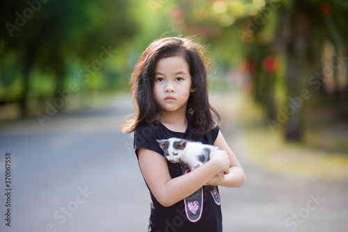 Asian young girl holding a cat kitten in the park under the sun. Pet and domestic animal lover. Happy child in nature during the summer. Friendship and childhood memory.