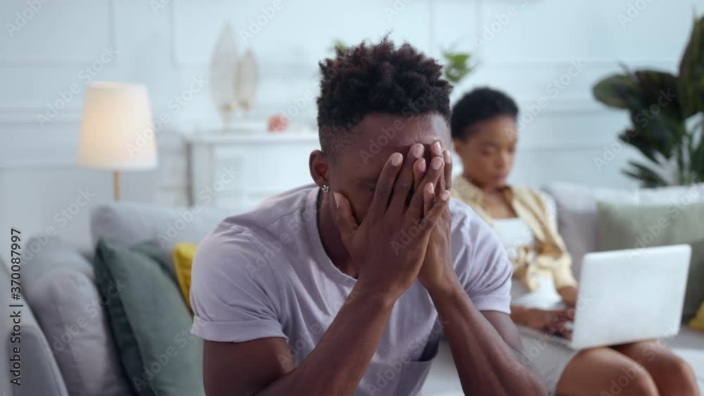 Sad irritated afro-american man husband sitting in despair worry about family problems and internet addiction. His indifferent wife sitting behind with laptop. Sad irritated afro-american man husband sitting in despair worry about family problems and internet addiction. His indifferent wife sitting behind with laptop.