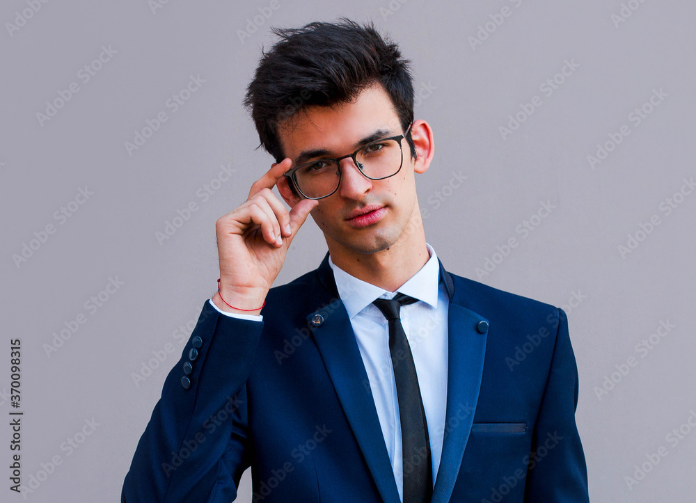 Young handsome man in blue suit and glasses smiling isolated on gray background Young handsome man in blue suit and glasses smiling isolated on gray background