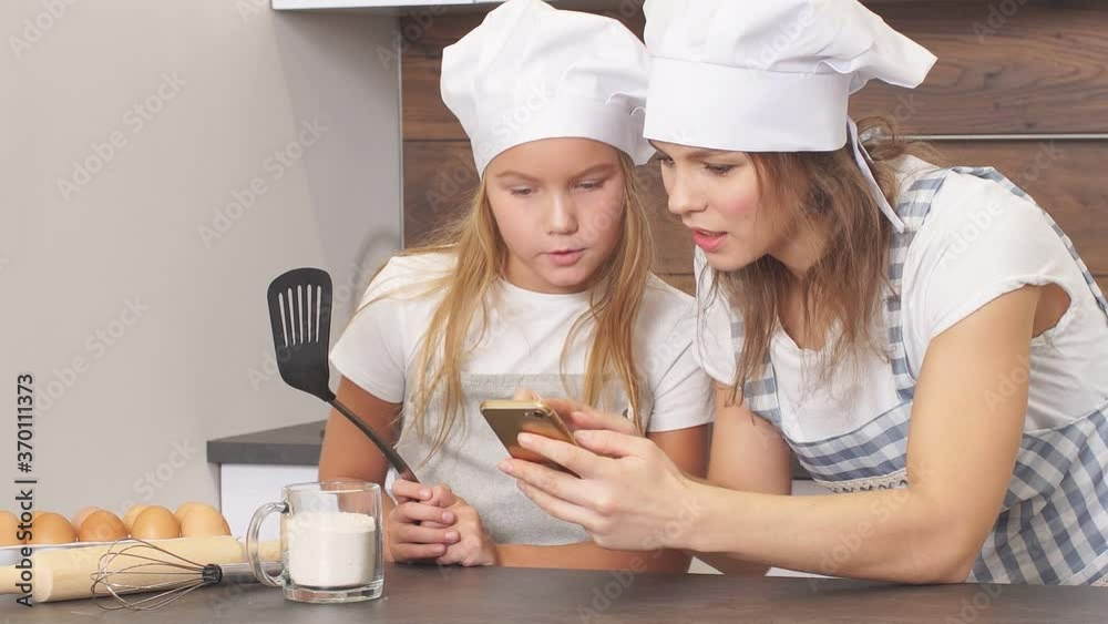 Happy family in aprons look at screen of smartphone watching delicious cake recipe, going to cook together Happy family in aprons look at screen of smartphone watching delicious cake recipe, going to cook together