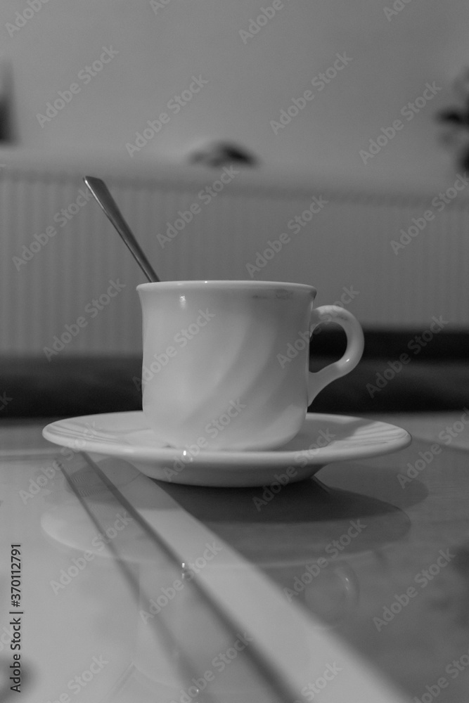 Small white ceramic coffee cup with a handle and a spoon placed on a white ceramic plate on a table Small white ceramic coffee cup with a handle and a spoon placed on a white ceramic plate on a table