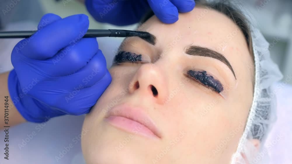 Cosmetologist making lash laminating, eyebrow and lash painting for woman in beauty clinic, face closeup. Beautician applying dark brown paint on woman's brows using brush in cosmetology. Cosmetologist making lash laminating, eyebrow and lash painting for woman in beauty clinic, face closeup. Beautician applying dark brown paint on woman's brows using brush in cosmetology.