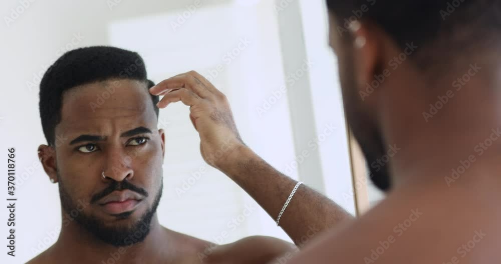 Focused young shirtless african ethnicity man looking in mirror, styling hair with fingers, getting ready in morning. Serious handsome naked mixed race guy doing hairstyle after morning showering. Focused young shirtless african ethnicity man looking in mirror, styling hair with fingers, getting ready in morning. Serious handsome naked mixed race guy doing hairstyle after morning showering.