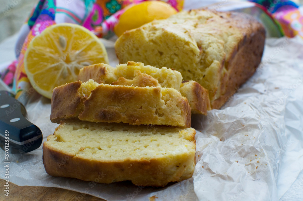 Homemade cake or baking concept. A vanilla or lemon loaf cake freshly baked and sliced on a wooden cutting board. There are lemons and a colourful tea towel in the background. Homemade cake or baking concept. A vanilla or lemon loaf cake freshly baked and sliced on a wooden cutting board. There are lemons and a colourful tea towel in the background.