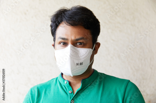 Adult Indian Male wearing a White N95 Respirator in Green T-Shirt facing and looking into camera (Black eyes, N95 Mask, White Backdrop, Natural Light, Mumbai, India, Protection of Illness)