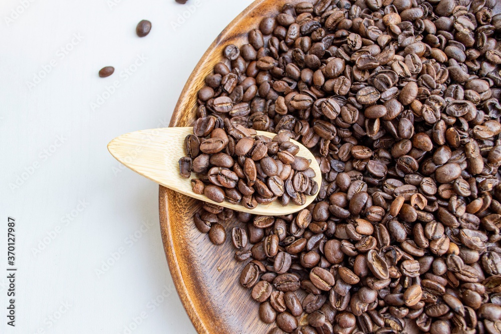 Fototapeta premium Composition in the rays of the bright sun. Beautifully roasted brown coffee beans on a round dark wood plate and on a small wooden spoon (selective focus not on all beans)