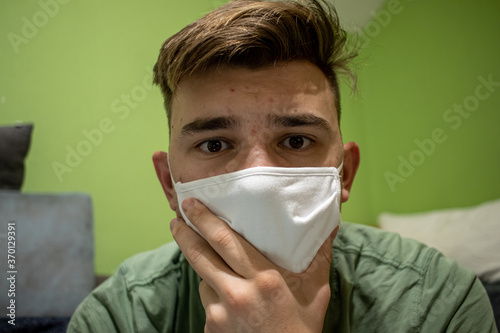 Young male with light brown hair wearing a white medical mask having a devastated and worried look in a green room