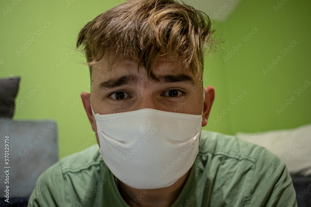 Young male with light brown hair wearing a white medical mask having a devastated and worried look in a green room Young male with light brown hair wearing a white medical mask having a devastated and worried look in a green room