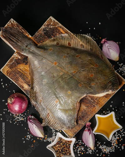 Top view turbot flounder on a wooden cutting board on a dark background. Red onion, salt, pepper and spices
