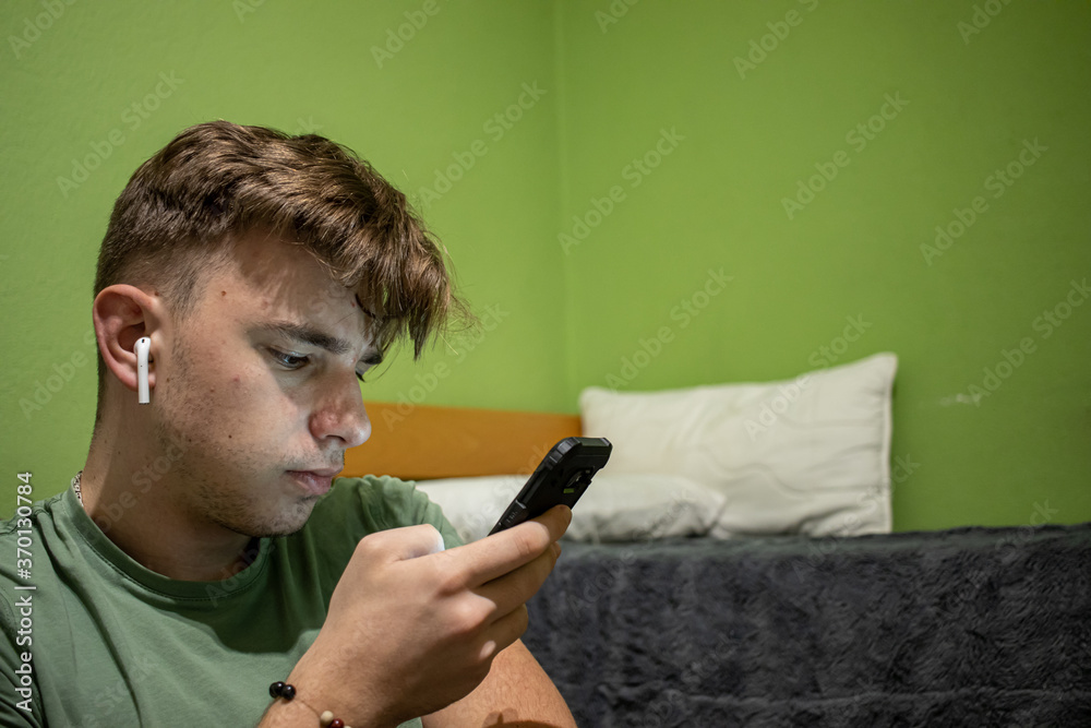 Young male with light brown hair using his black smartphone in a green bedroom Young male with light brown hair using his black smartphone in a green bedroom