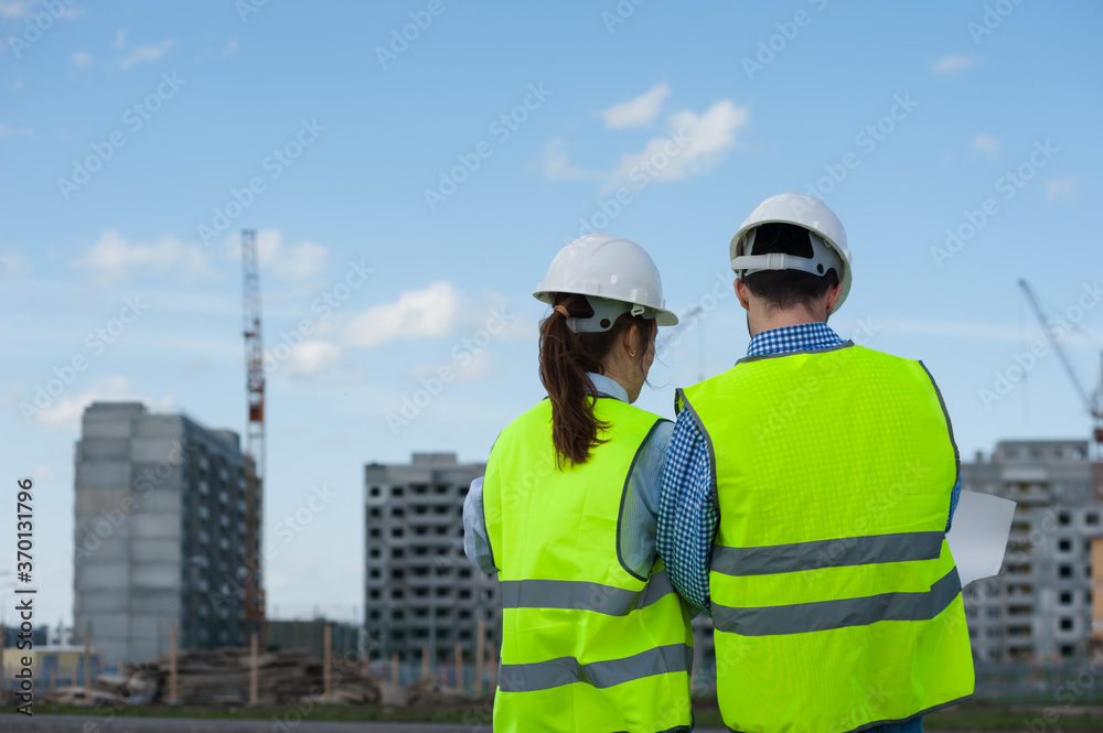 Rear View - Two Young Engineers A Man And A Woman In Green Vests And Helmets Are Discussing A Plan Rear View - Two Young Engineers A Man And A Woman In Green Vests And Helmets Are Discussing A Plan
