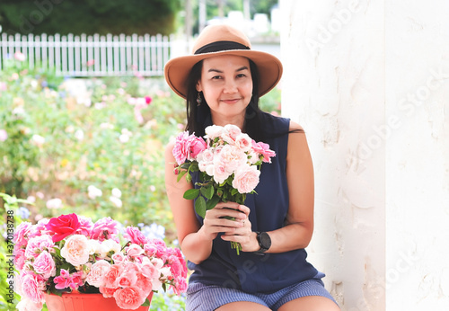 happy Asian woman wearing hat holding roses bouquet,sitting with rose bucket in her rose garden , smiling happily and looking at camera.