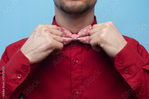 Leinwand Poster Close-up of the hands of a young man in a red shirt correcting bow-tie against a blue background