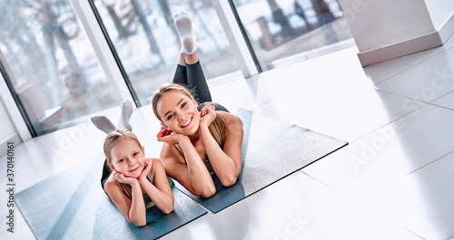 Beautiful young woman and charming little girl are looking at camera and smiling while lying on yoga mat in fitness hall © HBS