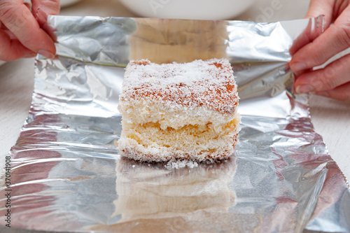 Traditional Brazilian dessert (known as Bolo Gelado) - Making step by step: Woman hand wrapping sliced cake sprinkled with granulated coconut in aluminum foil. Close-up. Horizontal shot. photo