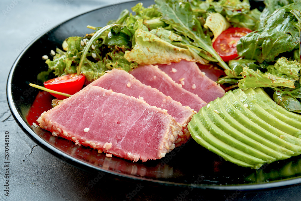 Salad with tuna, avocado and cherry tomatoes in a black plate on a gray background. Close up. Selective focus. Half of plate Salad with tuna, avocado and cherry tomatoes in a black plate on a gray background. Close up. Selective focus. Half of plate