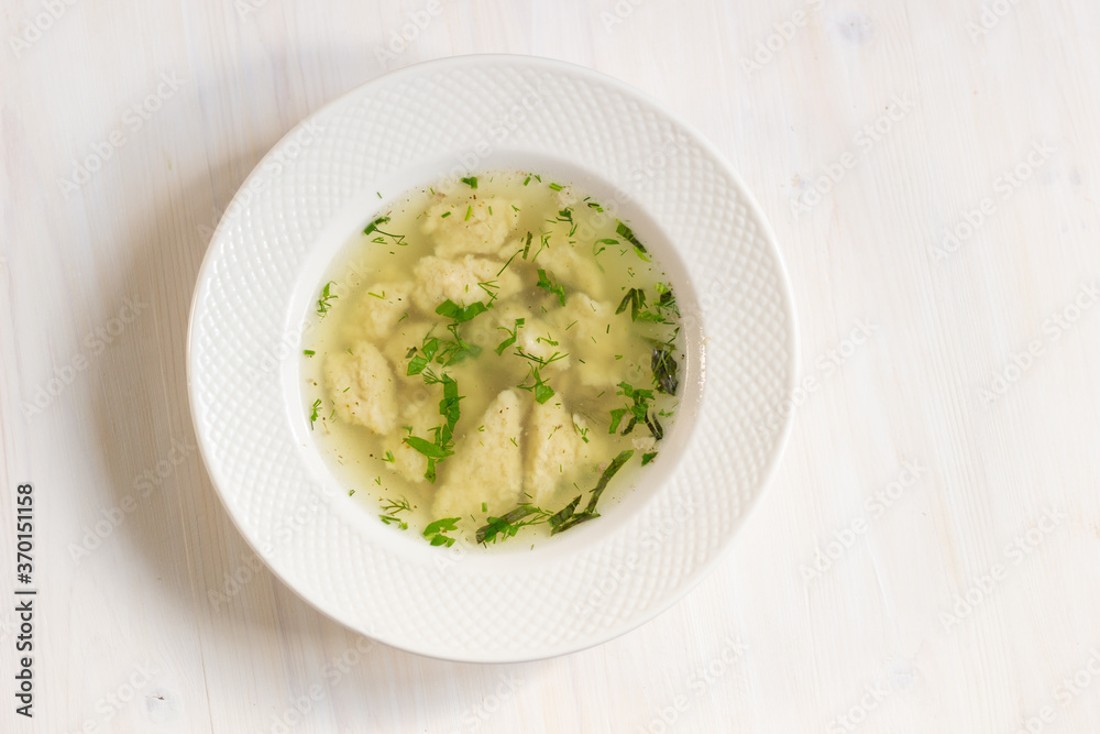 Chiken broth with dough and herbs in plate on white wooden background