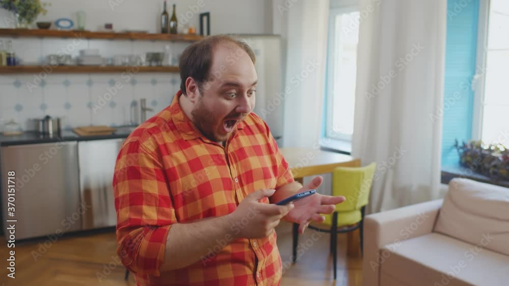 Excited obese young man using smartphone celebrating victory or winning lottery standing at home Excited obese young man using smartphone celebrating victory or winning lottery standing at home