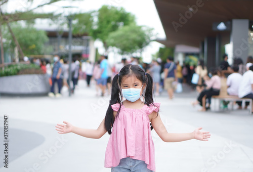 Asian child or kid girl smile wear paper face mask and open arm to present on lot of people for social distancing from person enjoy shopping at department store or market mall on holiday in covid era © kornnphoto