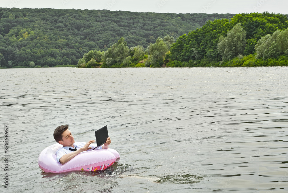 A man in a shirt on a pink inflatable circle. Guy with a laptop swims in the river. The concept of work during vacation, outsourcing and freelance. Place for text and copy space near businessman.