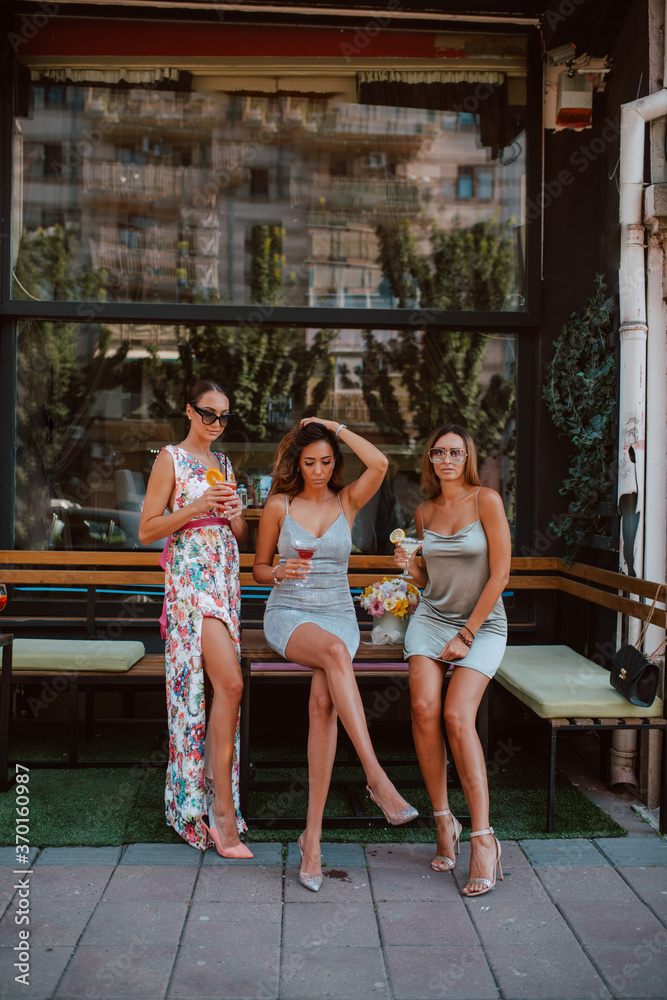 Three beautiful young caucasian women in short summer dresses are drinking cocktails in a cafe garden. Girls at party Three beautiful young caucasian women in short summer dresses are drinking cocktails in a cafe garden. Girls at party