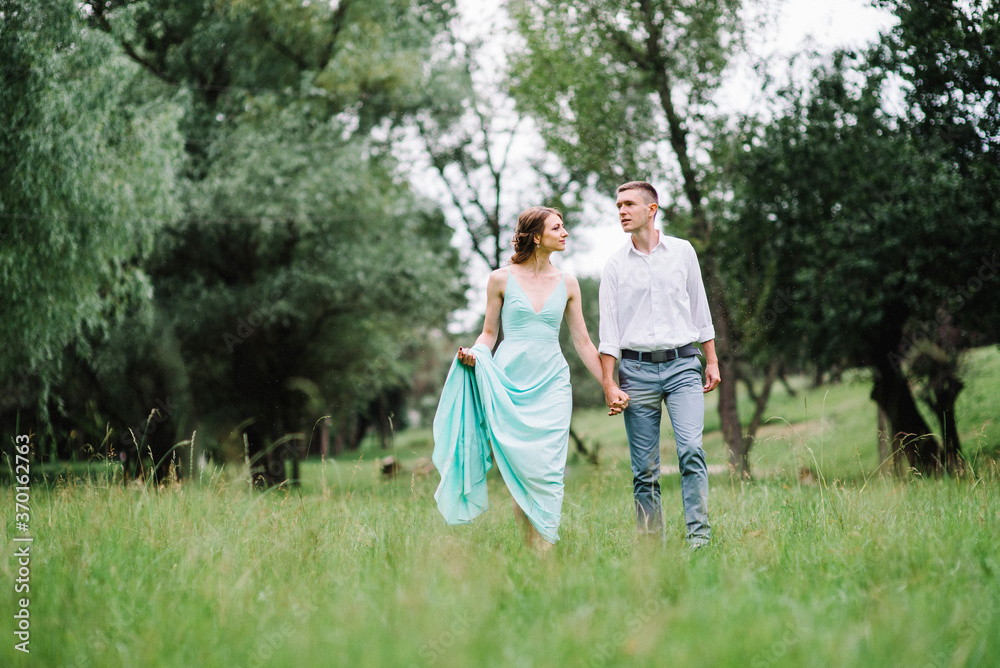 happy guy in a white shirt and a girl in a turquoise dress are walking in the forest park