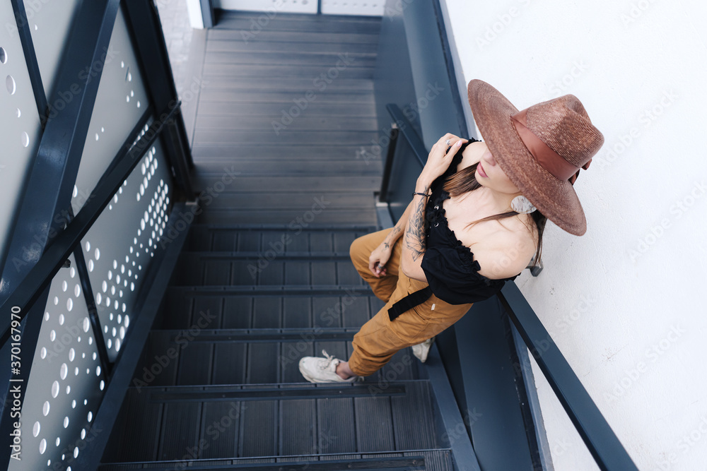 Obraz premium Fashionable brunette woman with long hair, wearing stylish large wicker hat, posing on the stairs. Young beautiful happy smiling fashionable traveller, tourist lady
