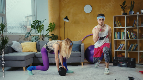 Funny weird fitness guy exercising lightly and dancing during home workout. His hardworking girlfriend trying to lift heavy dumbbells. Injustice. Sportive humor concept.
