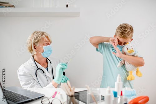 Pediatrician makes vaccination to cute Caucasian boy.They are wearing a protective face masks.