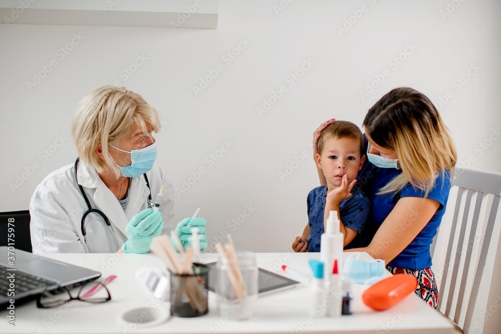 Pediatrician makes vaccination to cute Caucasian boy.Mother and doctor are wearing a protective face masks. Pediatrician makes vaccination to cute Caucasian boy.Mother and doctor are wearing a protective face masks.