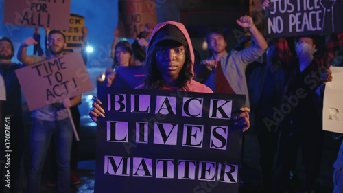Confident black woman holds invocatory poster, standing in crowd of shouting activists, protesting for equal rights and black lives matter. Solidarity demonstration.