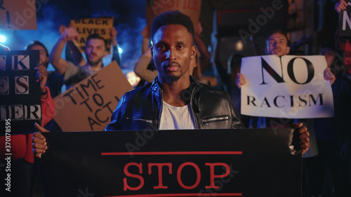 Fotografie Portrait black young man activist with Stop Racism banner protesting violence in multi-ethnic vigorous crowd on street riot