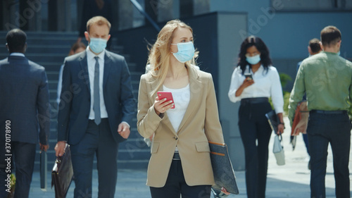 Blonde female emoloyer office manager businesswoman with face mask talking on smartphone walking home after work. Business district. Lockdown. Coronavirus. Self-isolation.
