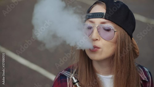 Close up pretty young hipster woman in black cap vape ecig. Girl vape popular ecig gadget, vaping device.Happy brunette vaper girl with e-cig. Portrait of smoker female model with electronic cigarette photo