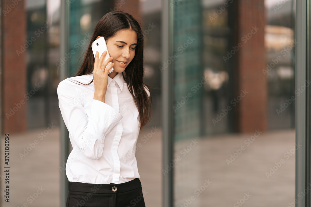 Fototapeta premium Portrait of a pretty Woman speaking on the phone wearing white business shirt. She is solving, negotiating daily tasks with her colleagues with glass walls in background.