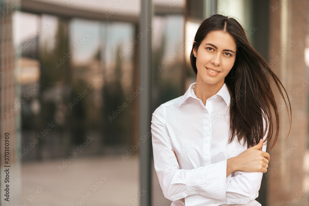 Portrait of a Caucasian pretty business lady smiling to the camera and crossing her hands while standing on the glass building background outdors Portrait of a Caucasian pretty business lady smiling to the camera and crossing her hands while standing on the glass building background outdors