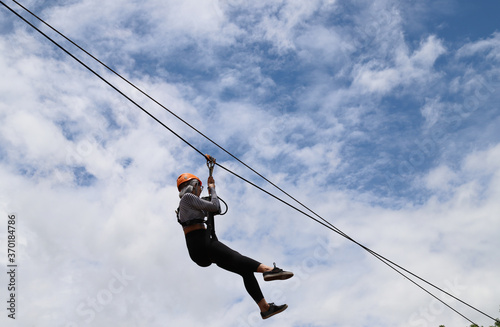 Closeup of young woman gliding on extreme trolley zip-line in adventure park with cloudy blue sky background. 