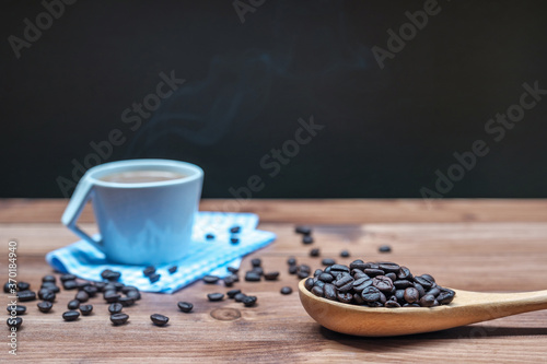 Roasted coffee beans on wood spoon and hot coffee in cup with smoke on the wood floor.