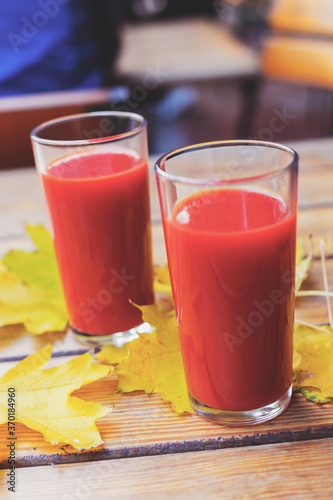 Tomato juice is poured into two glass glasses on a wooden table with yellow fallen maple leaves, in autumn. Selective focus. Tinted background