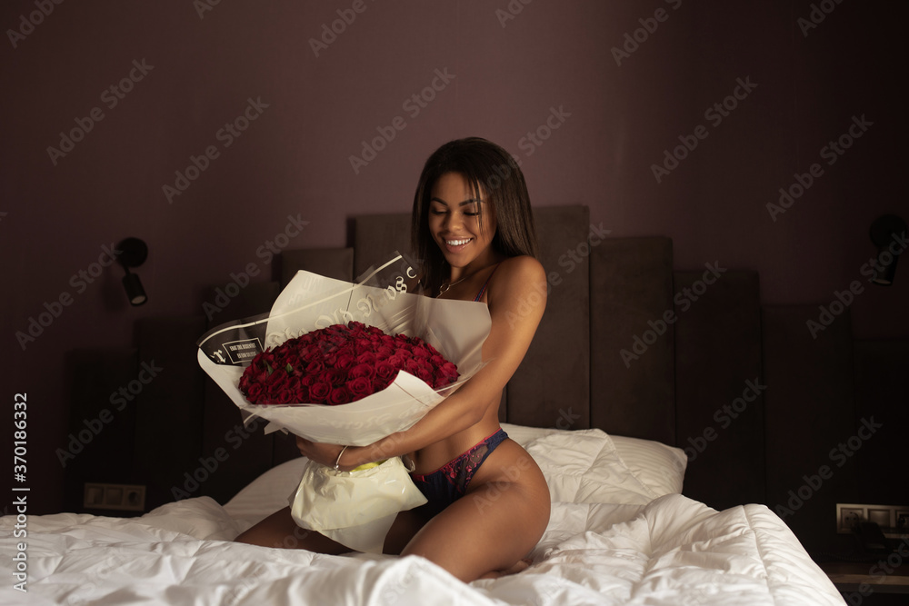 Young sexy black skin woman sitting on the bed and looking at bouquet of red roses.