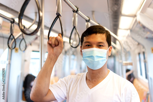 Young Asian man passenger wearing medical hygiene protective face mask are standing and holding handle ring inside subway or sky train in big city.