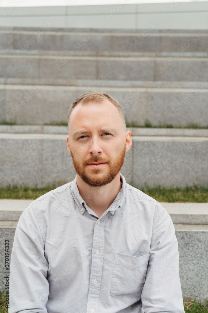 Portrait of a young man with a red beard and a fashionable haircut looking at the camera. A guy in a light shirt of casual style sits on a city street. Portrait of a young man with a red beard and a fashionable haircut looking at the camera. A guy in a light shirt of casual style sits on a city street.