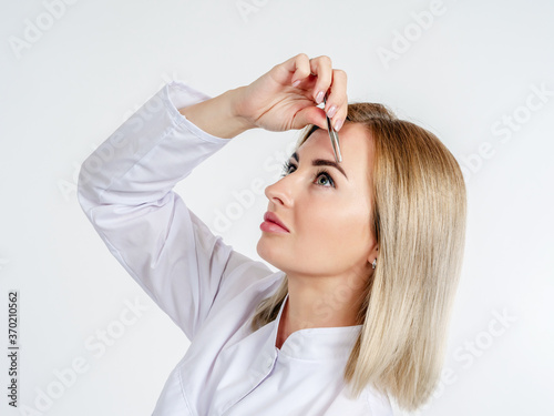 The girl plucks her eyebrows with tweezers. Eyebrow shaping. Eyebrow correction. A beautiful woman in white clothes adjusts her eyebrows.