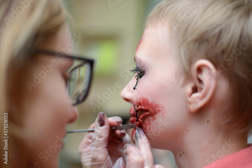 Make-up artist make the girl halloween make up in studio.Halloween face art.Woman applies on professional greasepaint on the face of blond girl.War paint with blood, scars and wounds. photo