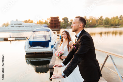 Beautiful luxurious couple, the groom in a black suit and the bride with a veil and a white wedding dress, holding hands, walk by the pool on the pier, next to the yacht and the ship © Олег Блохин