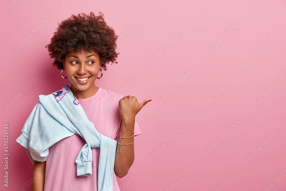 Pretty girl with Afro hair helps to pick best choice, points thumb aside on copy space, advertises product, smiles happily, wears rosy t shirt and sweater tied over shoulder. Your promo here