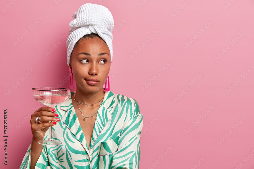 Photo of well groomed dark skinned woman holds glass of alcoholic beverage, looks aside, wears bath towel and dressing gown, thinks about something, has rest at home, poses against rosy background Photo of well groomed dark skinned woman holds glass of alcoholic beverage, looks aside, wears bath towel and dressing gown, thinks about something, has rest at home, poses against rosy background