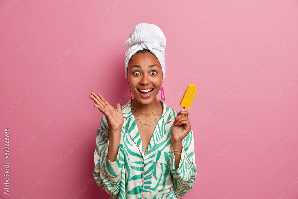 Summer, leisure and cold dessert. Smiling positive dark skinned woman holds delicious yellow mango ice cream on stick, feels excited and raises hand, wears towel wrapped on head after taking shower